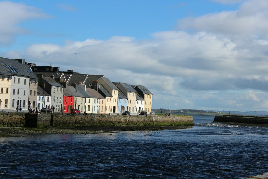 Galway Bay with its white and brown concrete building beside body of water under blue sky during daytime